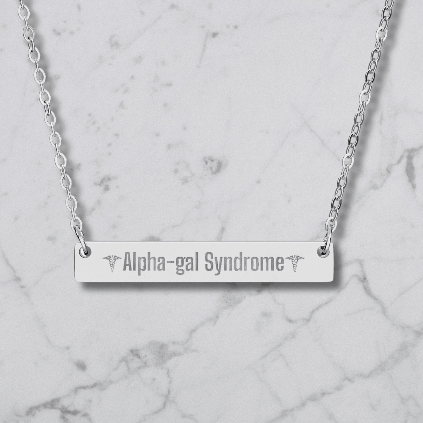 A silver bar necklace with the words 'Alpha-gal Syndrome' engraved in the center, flanked by caduceus symbols, displayed on a white marble background.