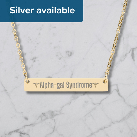 A gold bar necklace engraved with 'Alpha-gal Syndrome' and caduceus symbols, shown on a white marble background with a blue banner reading 'Silver available'.
