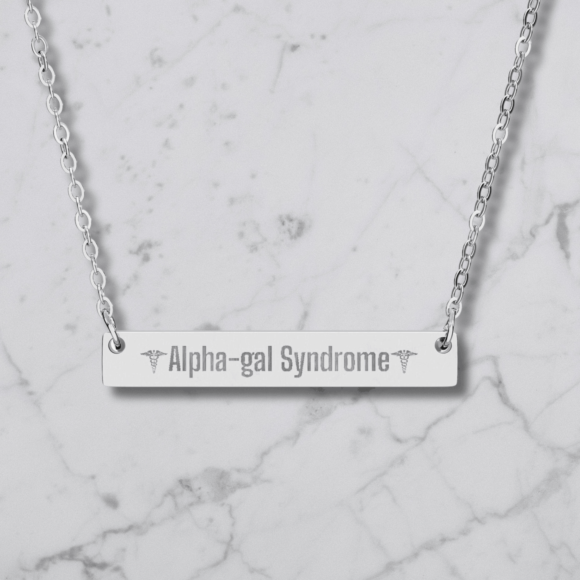 A silver bar necklace with the words 'Alpha-gal Syndrome' engraved in the center, flanked by caduceus symbols, displayed on a white marble background.
