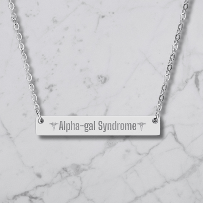 A silver bar necklace with the words 'Alpha-gal Syndrome' engraved in the center, flanked by caduceus symbols, displayed on a white marble background.